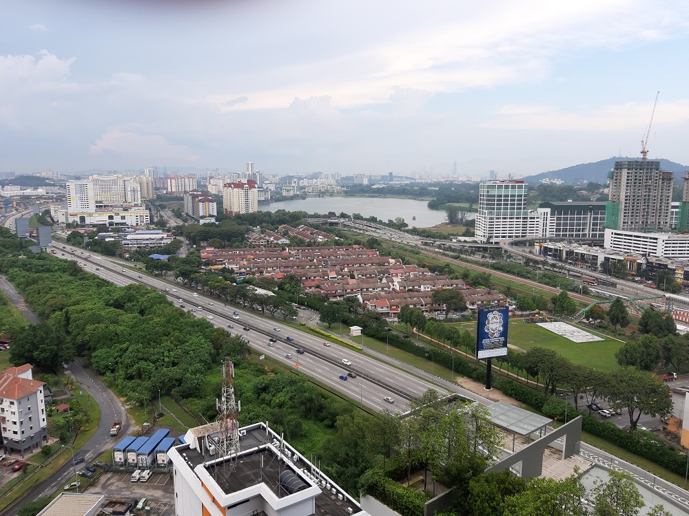 Daytime view from balcony showing highways, lake, green trees and cloudy sky
