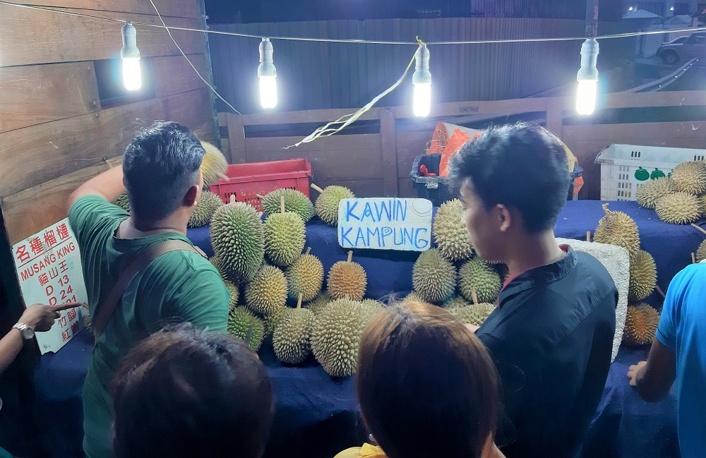 Durin stand at night lit by string of hanging lights as customers peruse the green fruits