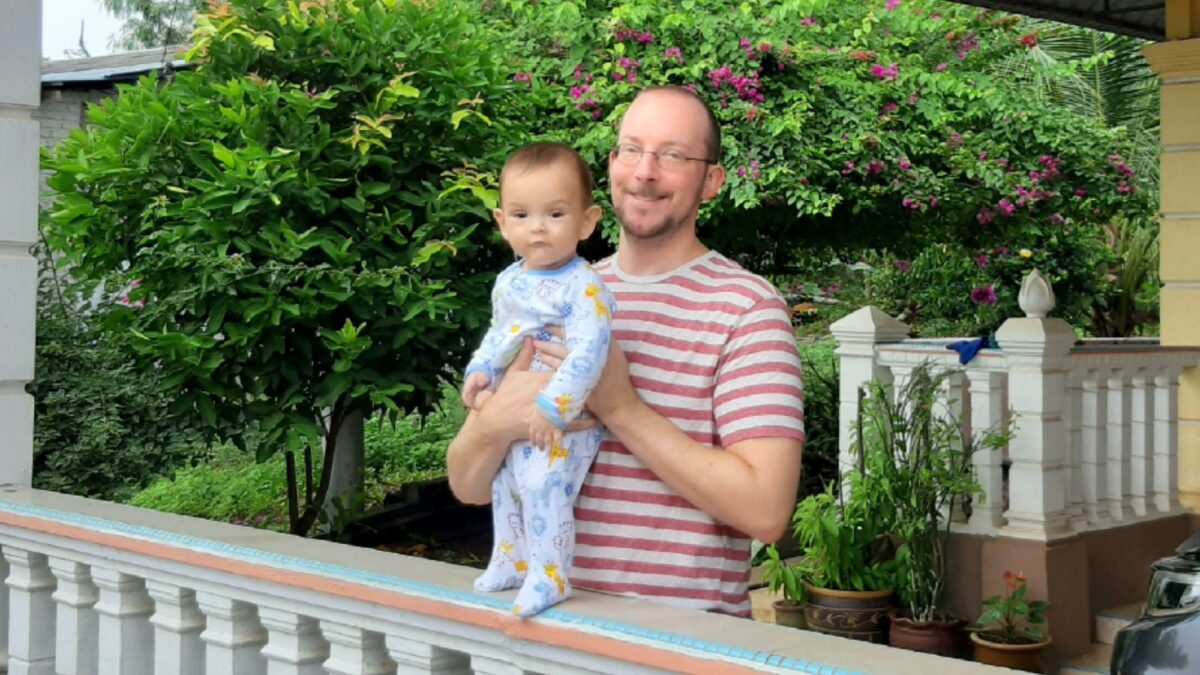 Father holding son outside on porch with flowering green tree in background