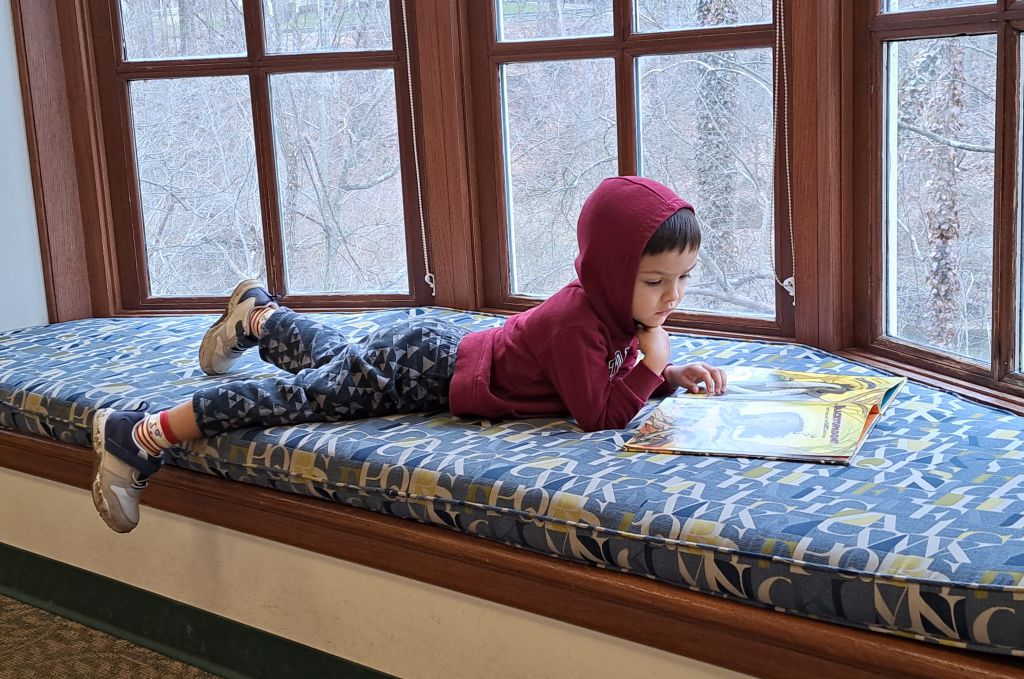 Adam lounging in a window nook reading a book and wearing a hoodie